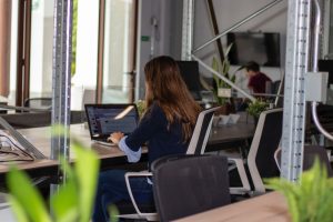 A woman sat at a desk in a modern office setting, typing on a laptop.