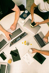 overhead shot of a group of people sat around a desk, all using smartphones and laptops together