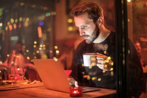 a man sits at a desk in a dimly lit room, sipping a coffee as he looks at a laptop