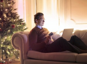 A man reclines on a sofa next to a Christmas tree. He has a credit card in hand and a laptop on his knees.