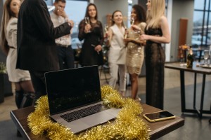 a laptop and phone sit abandoned on a desk, tinsel wrapped around them as people in formal wear gather in the background.