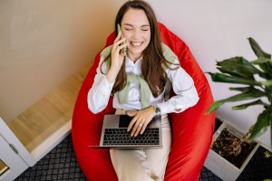 a woman sat in a red bean bag chair is on the phone and smiling broadly, as she works on a laptop