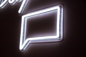 A white neon sign on a dark coloured wall, in the shape of the 'speech box chat' icon