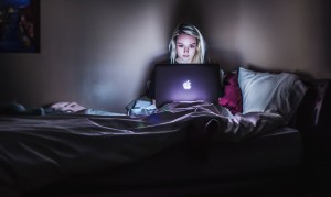 woman sat on a bed in a darkened room looking down at her Apple laptop, her face lit by the glow of the screen