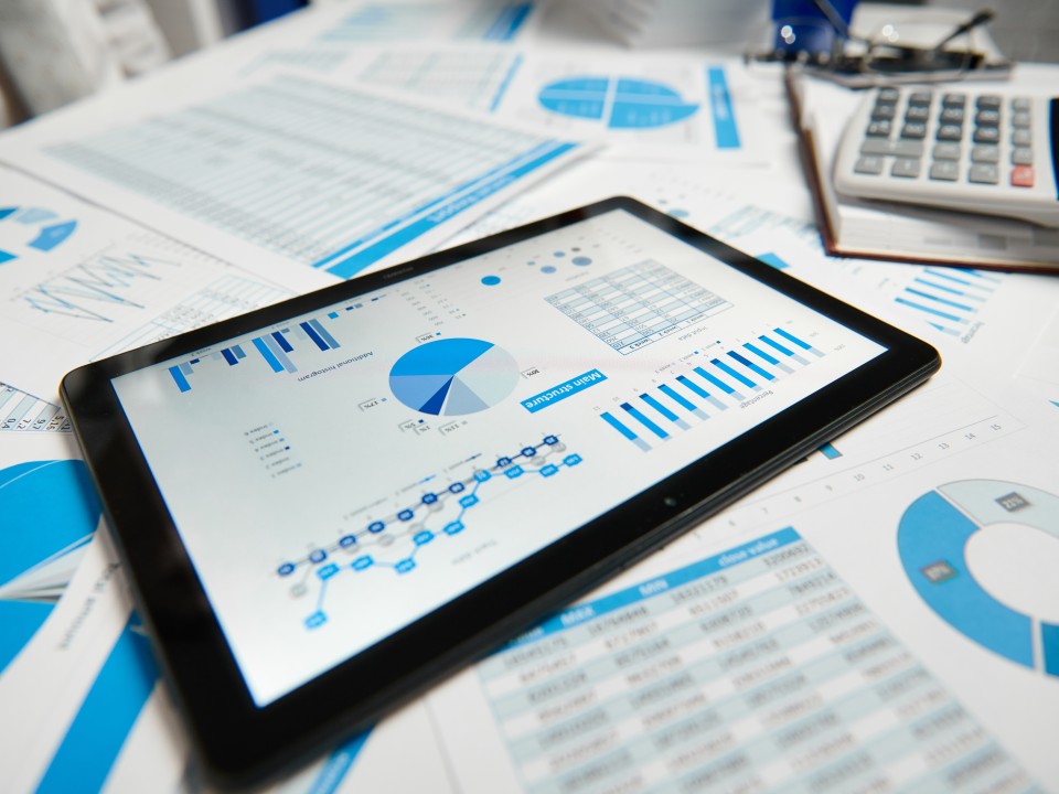 A table covered with lots of pieces of paper with various blue charts on, with a tablet laying on top showing more graphs and charts on it's screen. In the background, there is 2 pairs of glasses, and a calculator sat in the top right