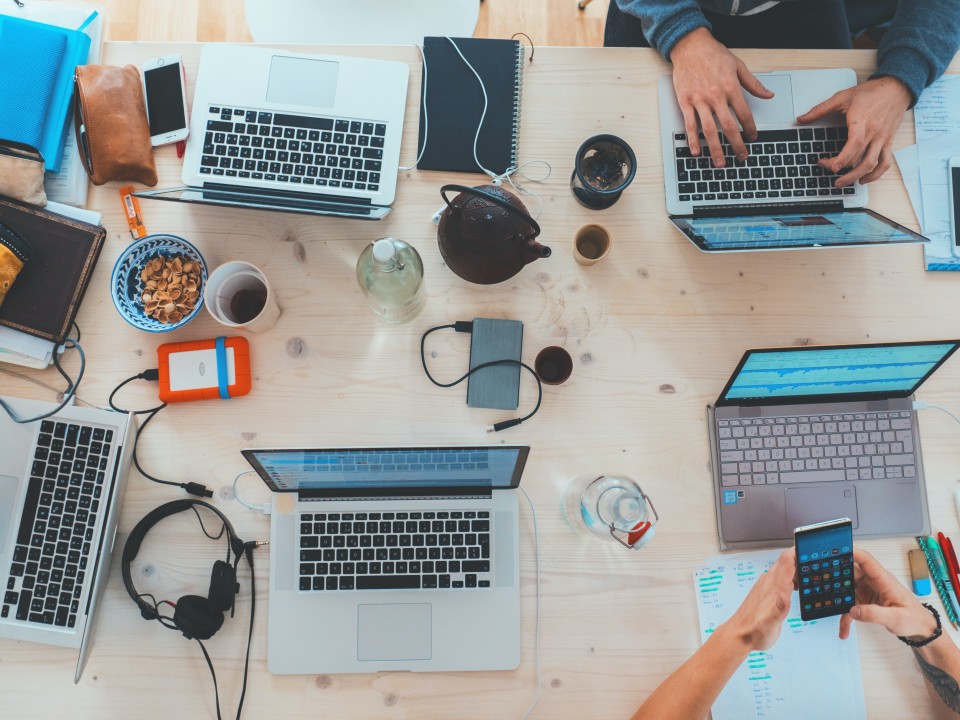 A top down view of a table. with at least 3 people areounf the table. The table is filled with laptops, pens, cups of coffee and notepads